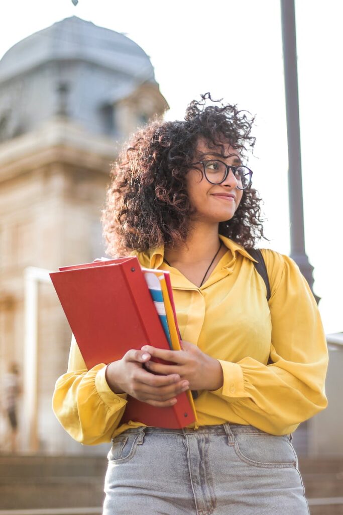 Smiling female student with curly hair and glasses holding books on campus steps.